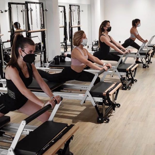 Women wearing masks performing Pilates on reformer machines in a studio.