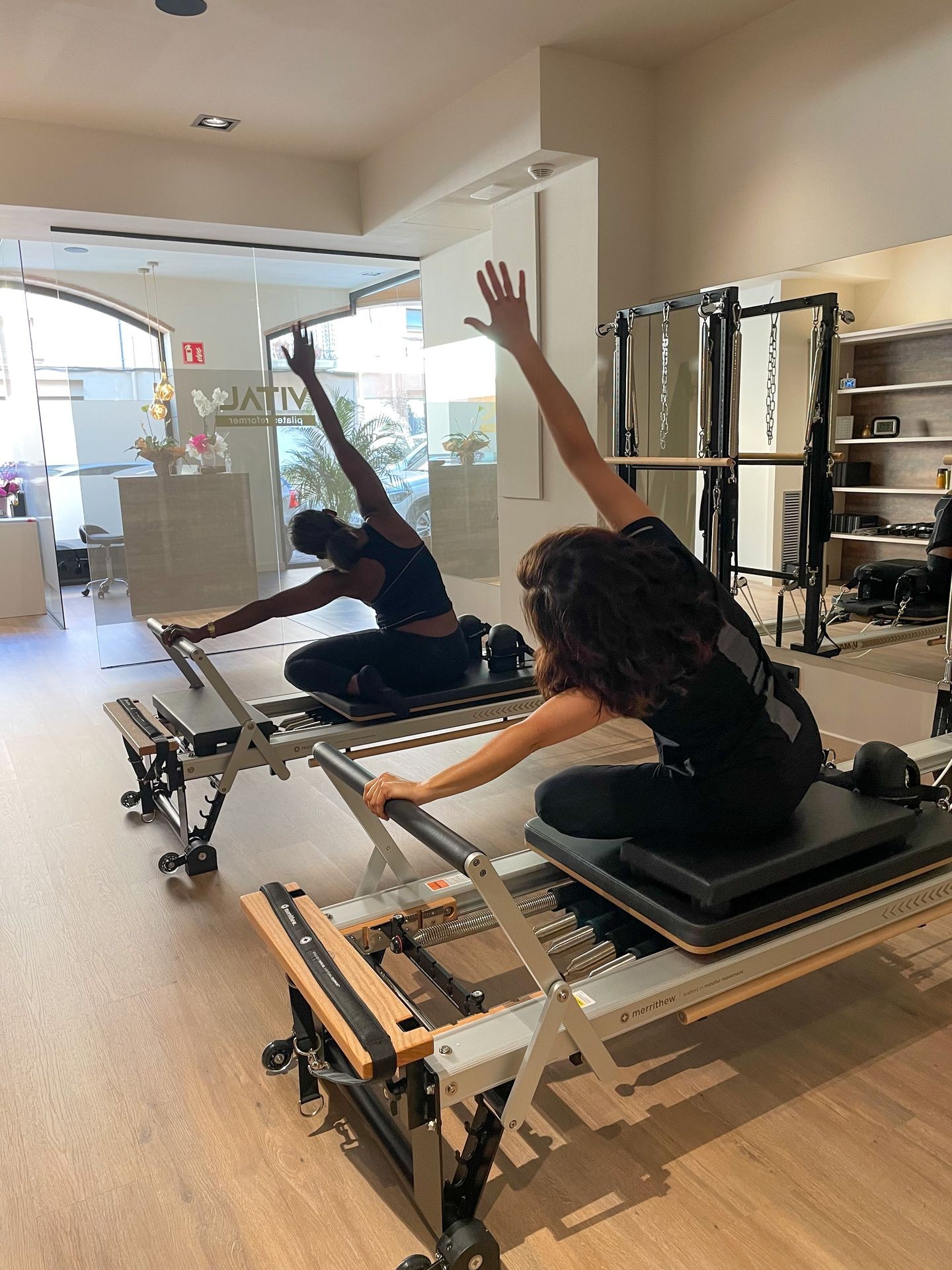 Two people on Pilates reformers stretching in a sunlit fitness studio.